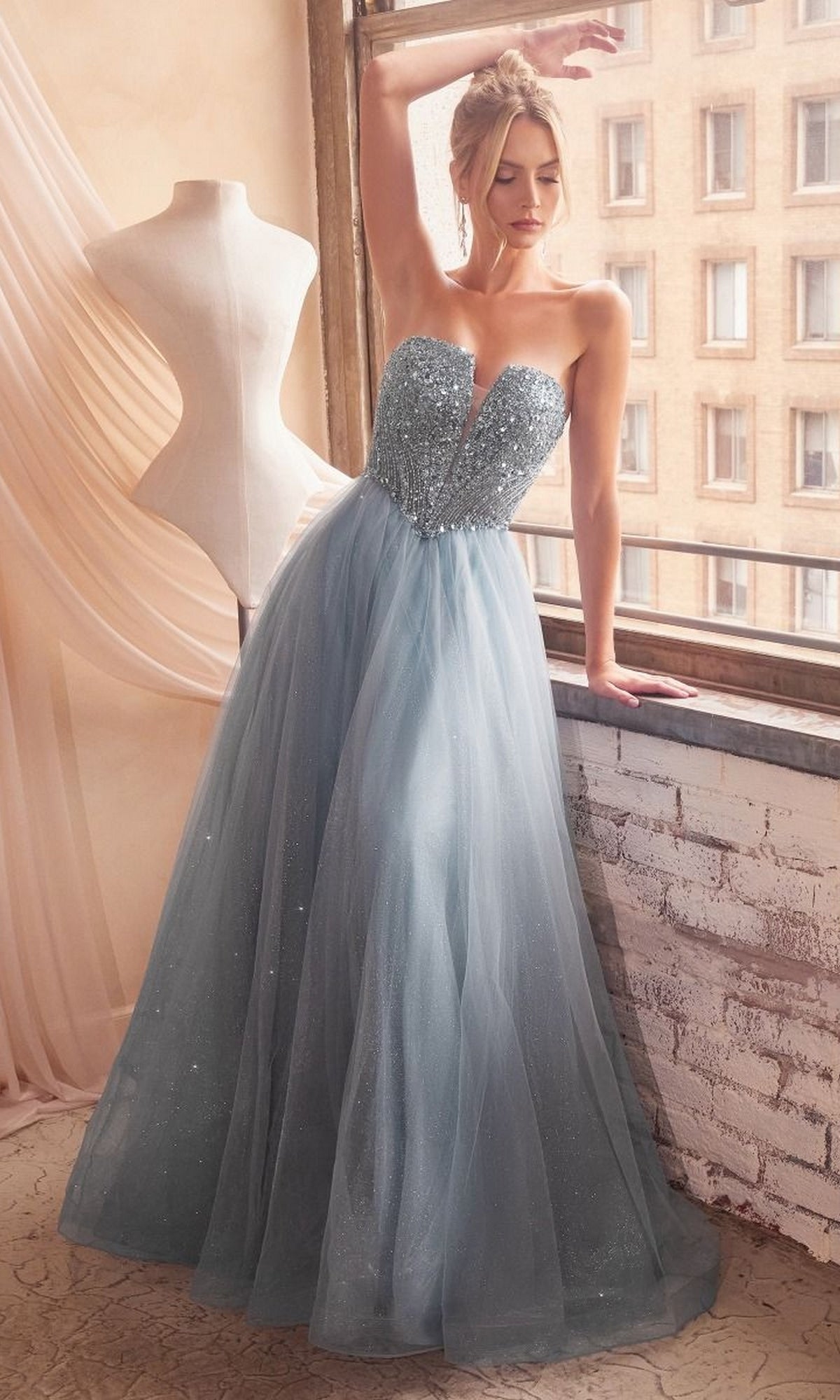 A woman in the Lace-Up Strapless Long Glitter Prom Dress CD0217 sparkles by the window, her shimmering light blue gown beside a dress form and sheer curtains.